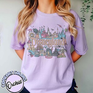 May include: A light purple Comfort Colors t-shirt featuring a colorful Disneyland Resort graphic. The graphic includes various park attractions and the Disneyland logo. The shirt has short sleeves and a crew neck.