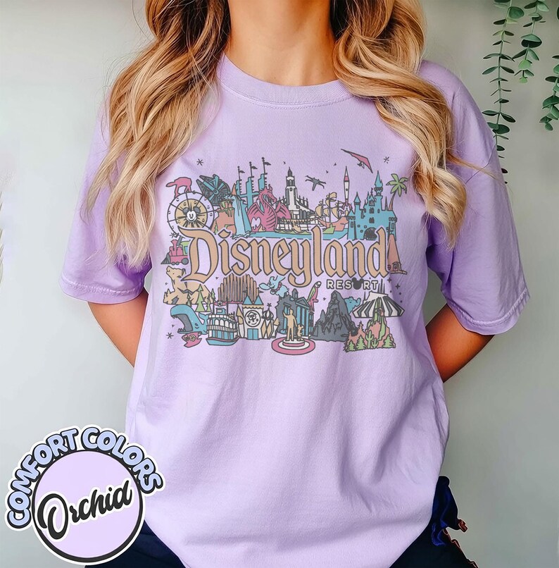 May include: A light purple Comfort Colors t-shirt featuring a colorful Disneyland Resort graphic. The graphic includes various park attractions and the Disneyland logo. The shirt has short sleeves and a crew neck.