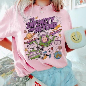 Puede incluir: Camiseta rosa claro con un dise&ntilde;o de Buzz Lightyear y la frase "To Infinity and Beyond". El dise&ntilde;o incluye planetas, un cohete y estrellas. Se sostiene una taza blanca con una carita sonriente amarilla.