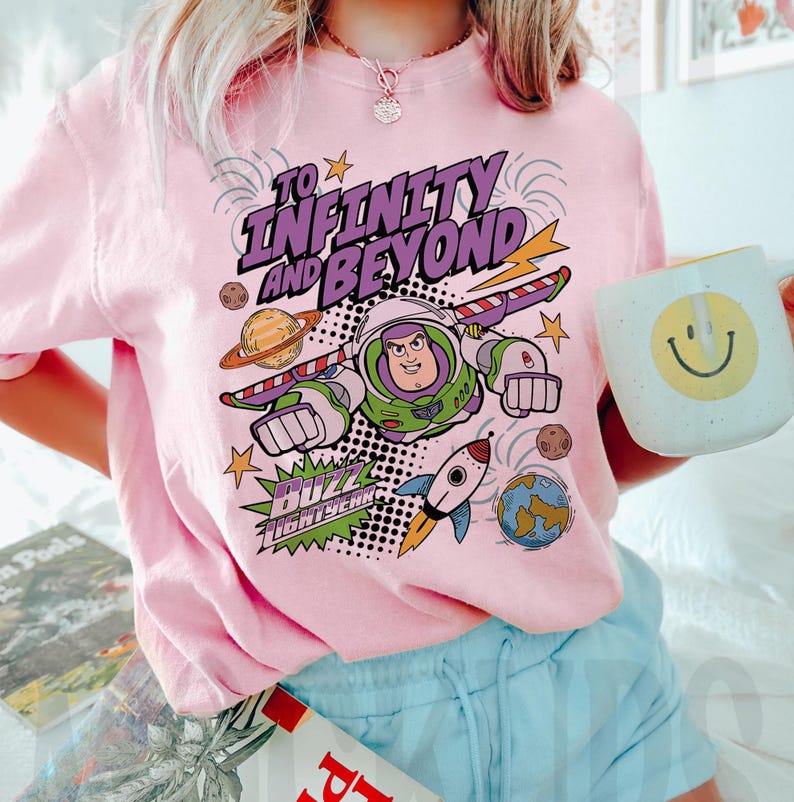 Puede incluir: Camiseta rosa claro con un dise&ntilde;o de Buzz Lightyear y la frase "To Infinity and Beyond". El dise&ntilde;o incluye planetas, un cohete y estrellas. Se sostiene una taza blanca con una carita sonriente amarilla.
