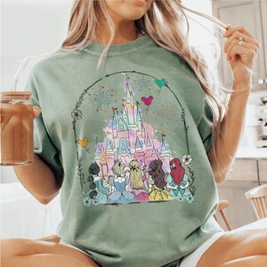 May include: A green t-shirt with a watercolor illustration of Cinderella's castle, fireworks, and seven Disney princesses. The princesses are wearing their signature dresses and are standing in front of the castle.
