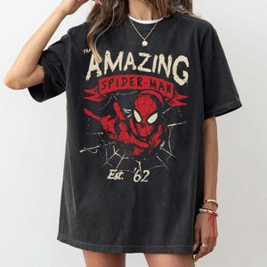 Pode incluir: Uma t-shirt de manga curta em preto carv&atilde;o com uma impress&atilde;o gr&aacute;fica do Spider-Man em vermelho e preto. O texto diz The Amazing Spider-Man e Est. 62, acompanhado por um design de teia de aranha. A t-shirt tem gola redonda e corte largo.