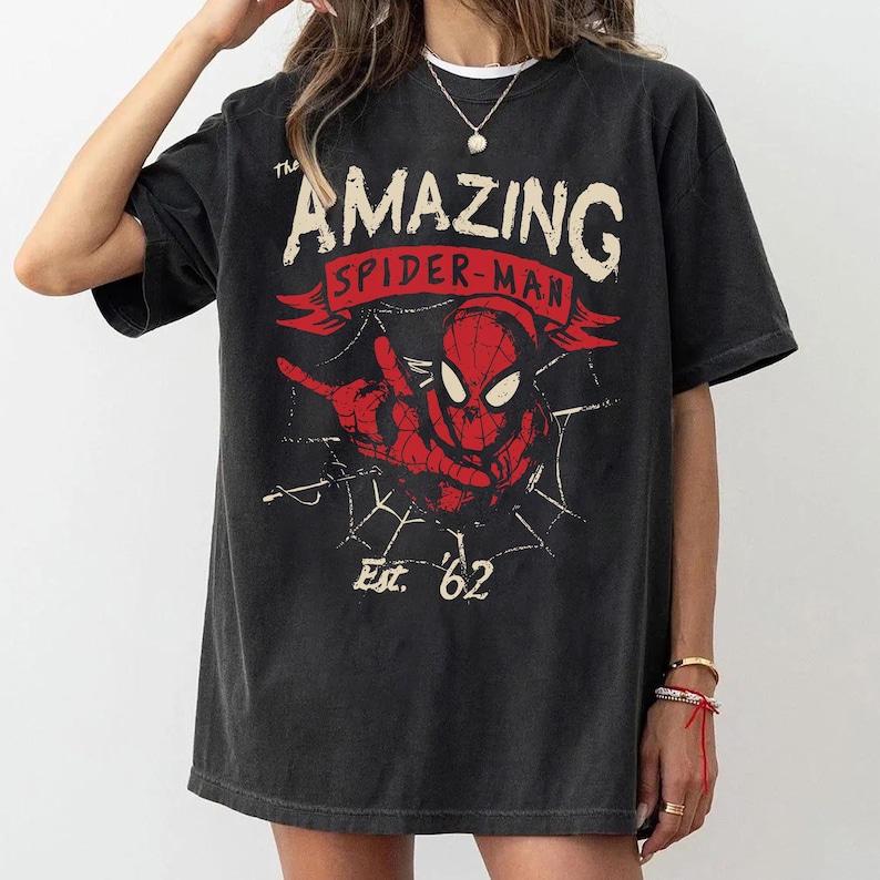 Pode incluir: Uma t-shirt de manga curta em preto carv&atilde;o com uma impress&atilde;o gr&aacute;fica do Spider-Man em vermelho e preto. O texto diz The Amazing Spider-Man e Est. 62, acompanhado por um design de teia de aranha. A t-shirt tem gola redonda e corte largo.
