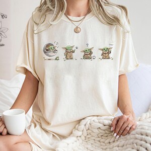 May include: A cream-colored short-sleeve t-shirt featuring a graphic print of a small green creature in four different poses. The illustrations show the character in a floating pod, using the force, looking confused with a question mark, and waving.