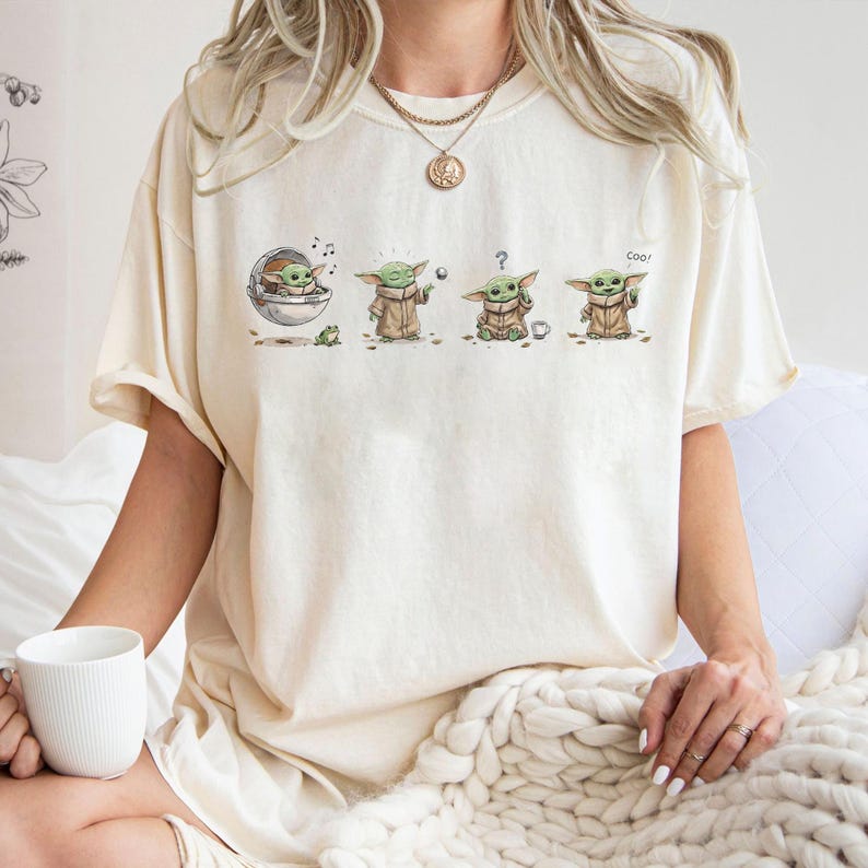 May include: A cream-colored short-sleeve t-shirt featuring a graphic print of a small green creature in four different poses. The illustrations show the character in a floating pod, using the force, looking confused with a question mark, and waving.