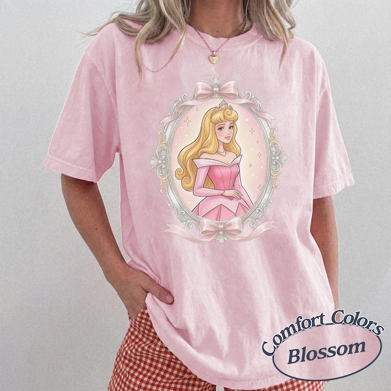 K&ouml;nnte beinhalten: Ein hellrosa T-Shirt mit kurzen &Auml;rmeln und einem zentralen Aufdruck einer blonden Figur in einem rosa Kleid, eingerahmt von einem silbernen Oval mit Schleifen oben und unten. Der Text Comfort Colors Blossom ist in der Ecke aufgedruckt.