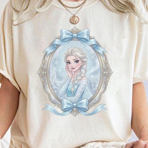 K&ouml;nnte beinhalten: Ein cremefarbenes T-Shirt mit einem Grafikdruck einer blonden Figur mit langem Zopf in einem blauen Kleid. Die Figur ist von einem verzierten ovalen Rahmen mit hellblauen Schleifen oben und unten vor einem verschneiten Hintergrund umgeben.