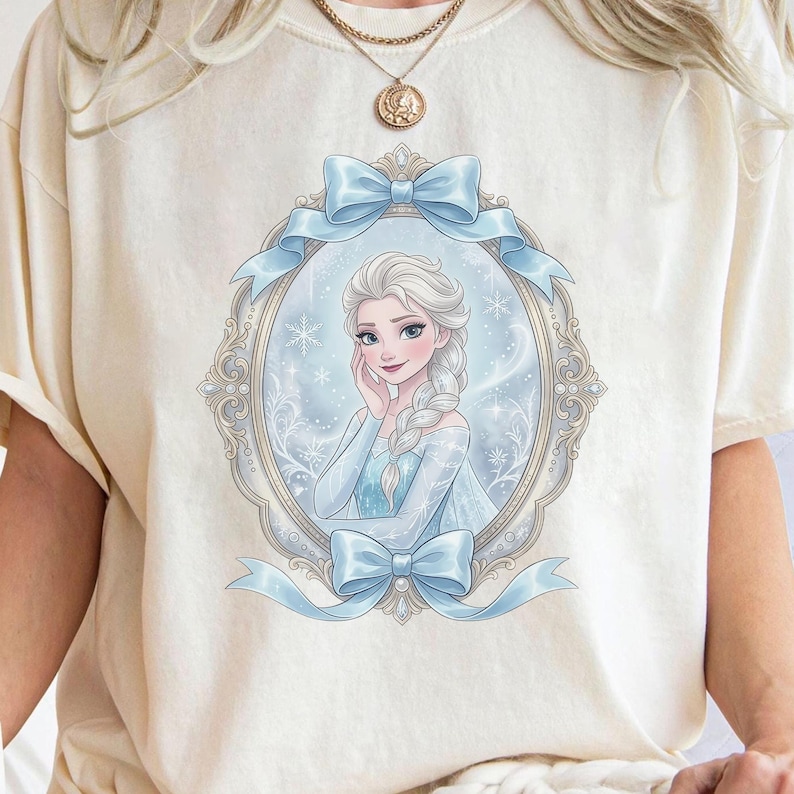 K&ouml;nnte beinhalten: Ein cremefarbenes T-Shirt mit einem Grafikdruck einer blonden Figur mit langem Zopf in einem blauen Kleid. Die Figur ist von einem verzierten ovalen Rahmen mit hellblauen Schleifen oben und unten vor einem verschneiten Hintergrund umgeben.