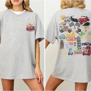 May include: A light gray t-shirt featuring a cartoon image of a red race car on the front and a collage of car-themed graphics on the back. The shirt has short sleeves and a crew neck.