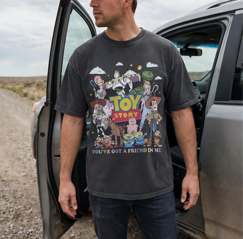 May include: Dark gray t-shirt featuring a colorful graphic of characters from the animated film "Toy Story." The shirt has the words "TOY STORY" in yellow and red, and the phrase "YOU'VE GOT A FRIEND IN ME" in white.