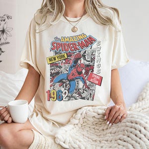 Peut inclure: T-shirt cr&egrave;me avec un graphisme de bande dessin&eacute;e vintage de Spider-Man. Le design comprend le texte "The Amazing Spider-Man", "New York" et "1962". Le super-h&eacute;ros est repr&eacute;sent&eacute; dans son costume rouge et bleu classique.
