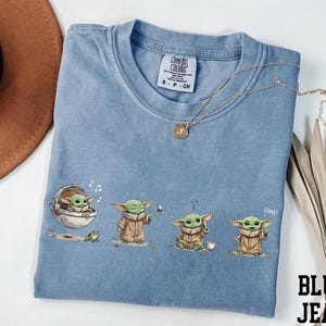 May include: A blue jean colored t-shirt featuring a graphic print of a small green creature in four different poses, including sitting in a floating pod, holding a silver ball, looking confused, and waving with the text coo! printed near the character.