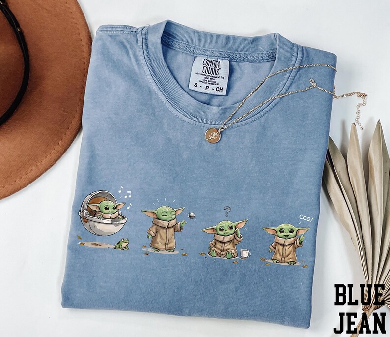 May include: A blue jean colored t-shirt featuring a graphic print of a small green creature in four different poses, including sitting in a floating pod, holding a silver ball, looking confused, and waving with the text coo! printed near the character.