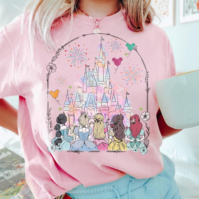 May include: A pink t-shirt with a graphic of Cinderella's castle, fireworks, and Disney princesses. The princesses are facing the castle and are wearing their signature dresses. The text "Disney" is not visible in the image.