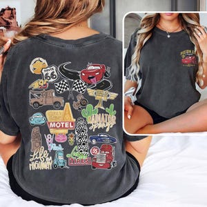 May include: Dark gray t-shirt featuring a colorful graphic of cartoon cars, Route 66, and other travel-themed elements. The back of the shirt has a large design, while the front pocket area has a smaller design with a red car and the text "Piston Cup".