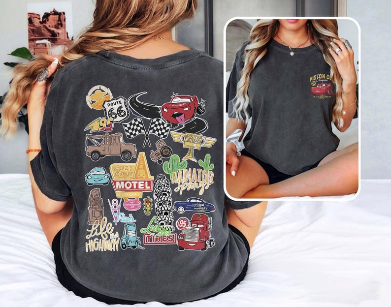 May include: Dark gray t-shirt featuring a colorful graphic of cartoon cars, Route 66, and other travel-themed elements. The back of the shirt has a large design, while the front pocket area has a smaller design with a red car and the text "Piston Cup".