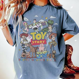 May include: A blue t-shirt featuring a colorful graphic of characters from the animated film "Toy Story." The design includes Woody, Buzz Lightyear, and other characters, with the words "Toy Story" and "You've Got a Friend in Me."