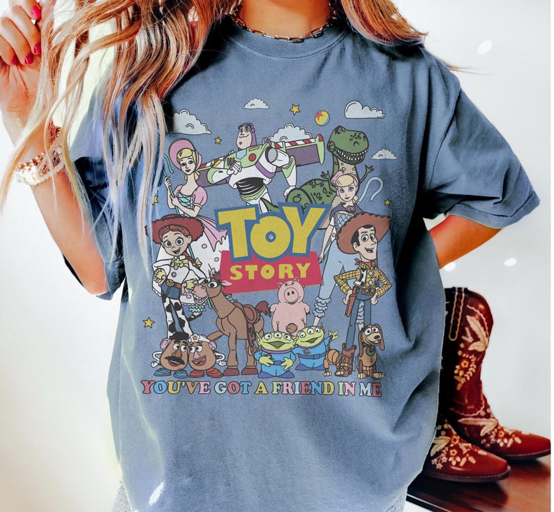 May include: A blue t-shirt featuring a colorful graphic of characters from the animated film "Toy Story." The design includes Woody, Buzz Lightyear, and other characters, with the words "Toy Story" and "You've Got a Friend in Me."