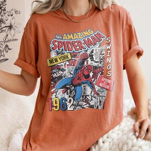 Peut inclure: T-shirt orange br&ucirc;l&eacute; avec un graphisme de bande dessin&eacute;e de Spider-Man. Le motif comprend le texte "The Amazing Spider-Man", "New York", "1962" et "Swings into Action". Le t-shirt a des manches courtes et une coupe d&eacute;contract&eacute;e.