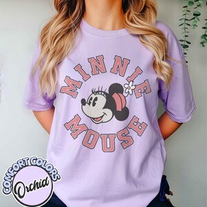 Puede incluir: Una camiseta lila con un dise&ntilde;o de Minnie Mouse. El dise&ntilde;o incluye las palabras "MINNIE MOUSE" en un c&iacute;rculo alrededor de la cara de Minnie. La camiseta tiene mangas cortas y cuello redondo. El logo "Comfort Colors" es visible.