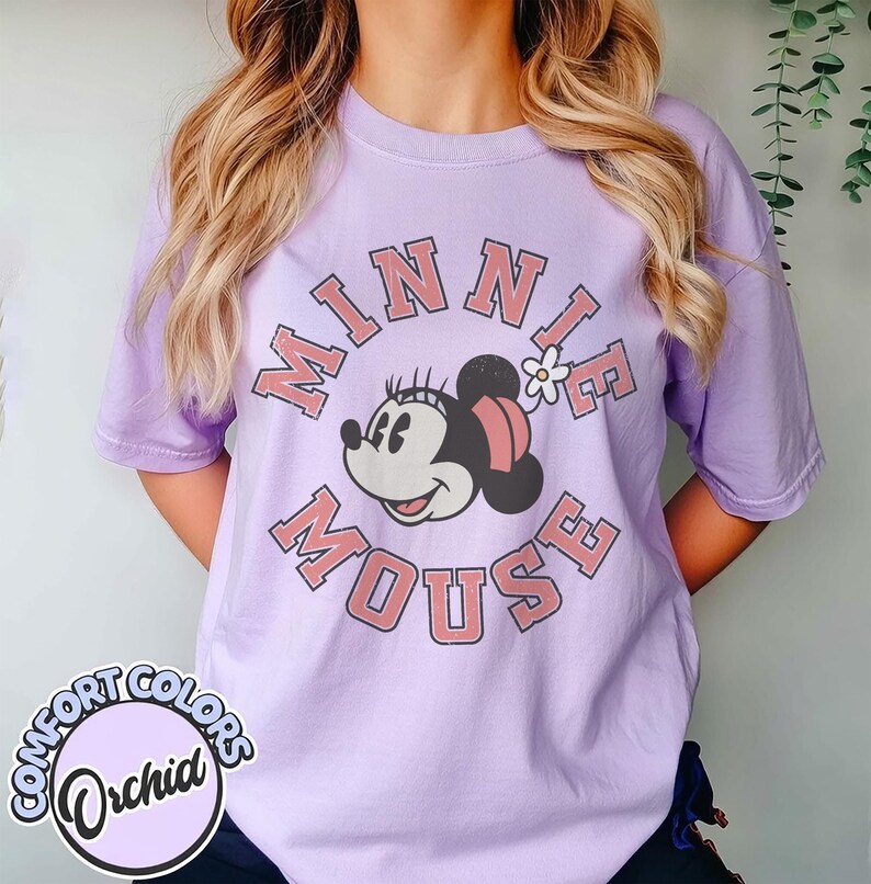 Puede incluir: Una camiseta lila con un dise&ntilde;o de Minnie Mouse. El dise&ntilde;o incluye las palabras "MINNIE MOUSE" en un c&iacute;rculo alrededor de la cara de Minnie. La camiseta tiene mangas cortas y cuello redondo. El logo "Comfort Colors" es visible.