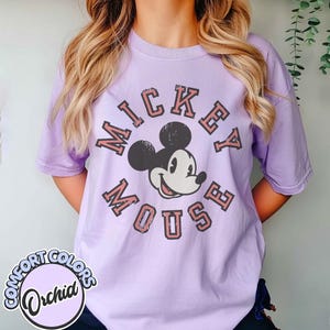 May include: A light purple t-shirt featuring a graphic of Mickey Mouse with the words "Mickey Mouse" in a vintage style. The shirt also has the Comfort Colors logo in a circle at the bottom.