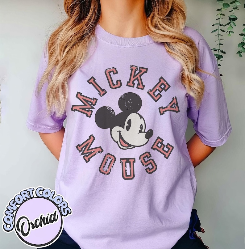 May include: A light purple t-shirt featuring a graphic of Mickey Mouse with the words "Mickey Mouse" in a vintage style. The shirt also has the Comfort Colors logo in a circle at the bottom.
