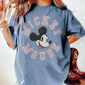 May include: A blue t-shirt featuring a distressed Mickey Mouse graphic. The design includes the words "Mickey Mouse" in a circular arrangement around Mickey's face. The shirt has short sleeves and a relaxed fit.