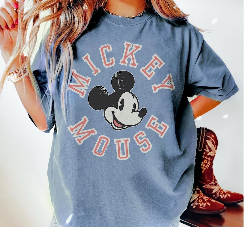 May include: A blue t-shirt featuring a distressed Mickey Mouse graphic. The design includes the words "Mickey Mouse" in a circular arrangement around Mickey's face. The shirt has short sleeves and a relaxed fit.