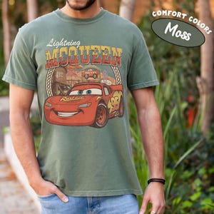 Puede incluir: Una camiseta de manga corta color verde musgo con un gr&aacute;fico del coche de carreras rojo Lightning McQueen. El dise&ntilde;o incluye el texto Lightning McQueen, el n&uacute;mero 95 y un fondo de desierto. La camiseta est&aacute; etiquetada como Comfort Colors Moss.