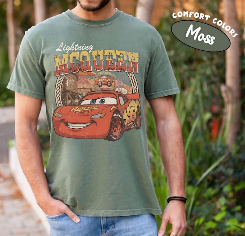 Puede incluir: Una camiseta de manga corta color verde musgo con un gr&aacute;fico del coche de carreras rojo Lightning McQueen. El dise&ntilde;o incluye el texto Lightning McQueen, el n&uacute;mero 95 y un fondo de desierto. La camiseta est&aacute; etiquetada como Comfort Colors Moss.