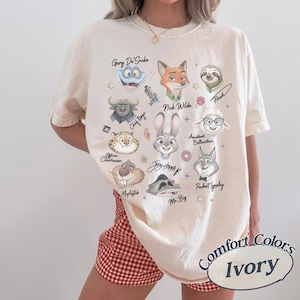 May include: An ivory short-sleeve t-shirt featuring a graphic print of various Zootopia characters including Judy Hopps, Nick Wilde, and Flash. The design includes character names in cursive script, small star icons, and a donut illustration.