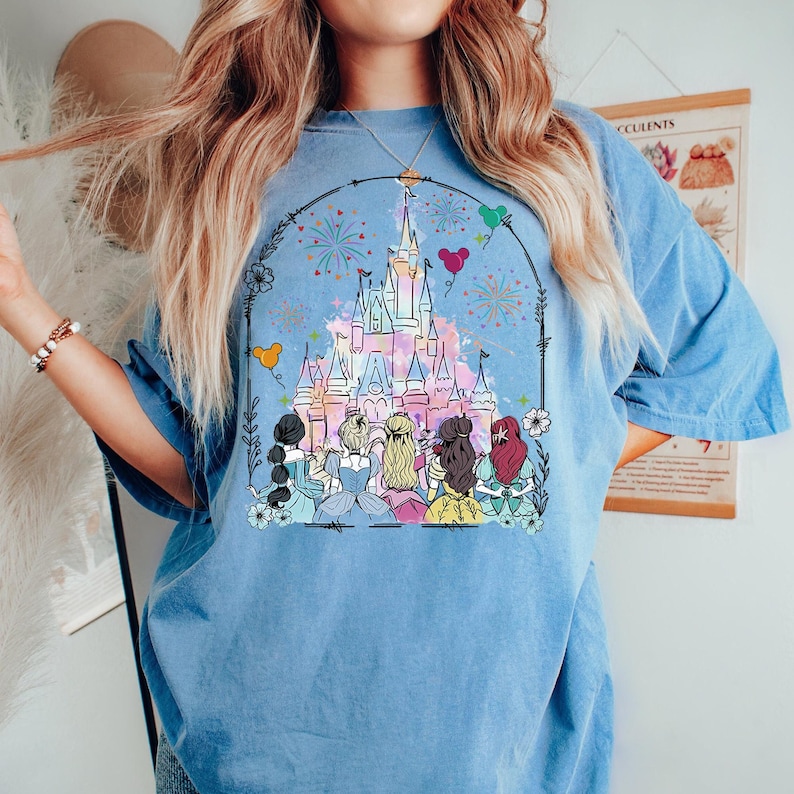 May include: A light blue t-shirt with a graphic of a pink and blue castle with fireworks and balloons. Seven princesses are standing in front of the castle.