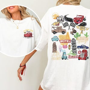 May include: White t-shirt featuring a graphic of a red race car with the text "Piston Cup '95 Champion" on the front. The back of the shirt has various cartoon car-themed illustrations, including Route 66 and checkered flags.