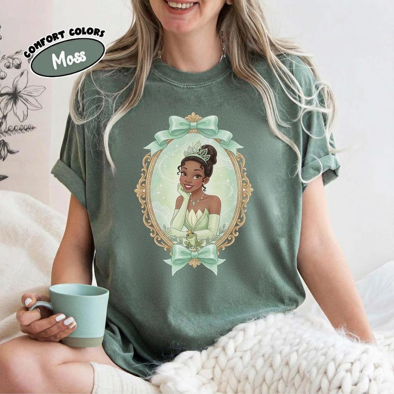 K&ouml;nnte beinhalten: Ein moosgr&uuml;nes Kurzarm-T-Shirt mit einer zentralen Grafik einer Figur, die eine Krone und ein hellgr&uuml;nes Kleid tr&auml;gt, platziert in einem verzierten goldenen ovalen Rahmen mit hellgr&uuml;nen Schleifen. Der Text COMFORT COLORS MOSS ist sichtbar.