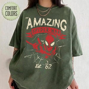 Pode incluir: Uma t-shirt de manga curta verde-azeitona com um gr&aacute;fico desgastado de uma m&aacute;scara vermelha do Spider-Man e linhas de teia de aranha brancas. O texto diz The AMAZING SPIDER-MAN e Est. 62. Um log&oacute;tipo circular Comfort Colors &eacute; vis&iacute;vel.