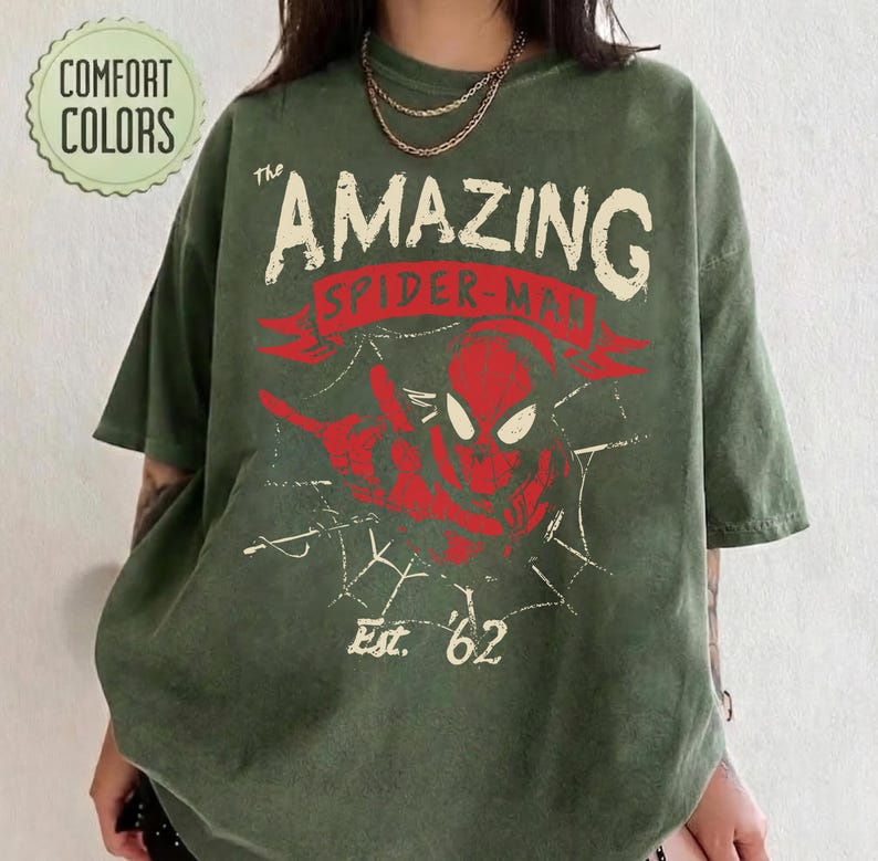 Pode incluir: Uma t-shirt de manga curta verde-azeitona com um gr&aacute;fico desgastado de uma m&aacute;scara vermelha do Spider-Man e linhas de teia de aranha brancas. O texto diz The AMAZING SPIDER-MAN e Est. 62. Um log&oacute;tipo circular Comfort Colors &eacute; vis&iacute;vel.