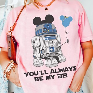 May include: Pink t-shirt featuring a cartoon R2-D2 character with Mickey Mouse ears and a blue balloon. The text "YOU'LL ALWAYS BE MY BB" is printed in black. The shirt is a light pink color.