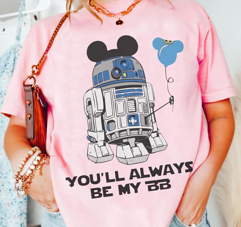 May include: Pink t-shirt featuring a cartoon R2-D2 character with Mickey Mouse ears and a blue balloon. The text "YOU'LL ALWAYS BE MY BB" is printed in black. The shirt is a light pink color.