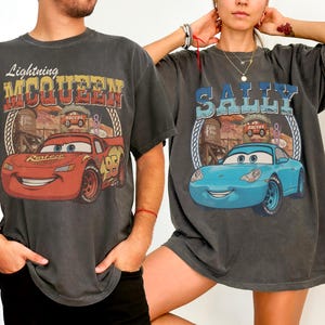 Puede incluir: Dos camisetas gris carb&oacute;n con estampados gr&aacute;ficos. La camiseta de la izquierda muestra un coche de carreras rojo con el texto Lightning MCQUEEN. La de la derecha muestra un coche azul claro con el texto SALLY. Ambos incluyen paisajes des&eacute;rticos.