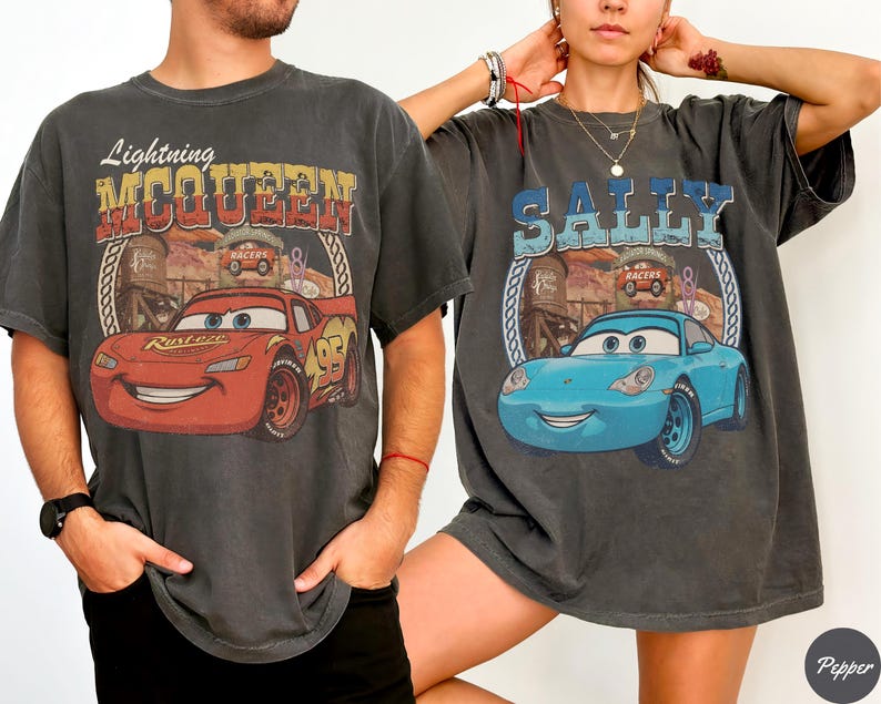 Puede incluir: Dos camisetas gris carb&oacute;n con estampados gr&aacute;ficos. La camiseta de la izquierda muestra un coche de carreras rojo con el texto Lightning MCQUEEN. La de la derecha muestra un coche azul claro con el texto SALLY. Ambos incluyen paisajes des&eacute;rticos.