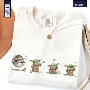 May include: An ivory t-shirt featuring a graphic print of a small green creature in four different poses. The print shows the character in a floating pod, using the force, looking confused with a question mark, and waving with the text Coo.