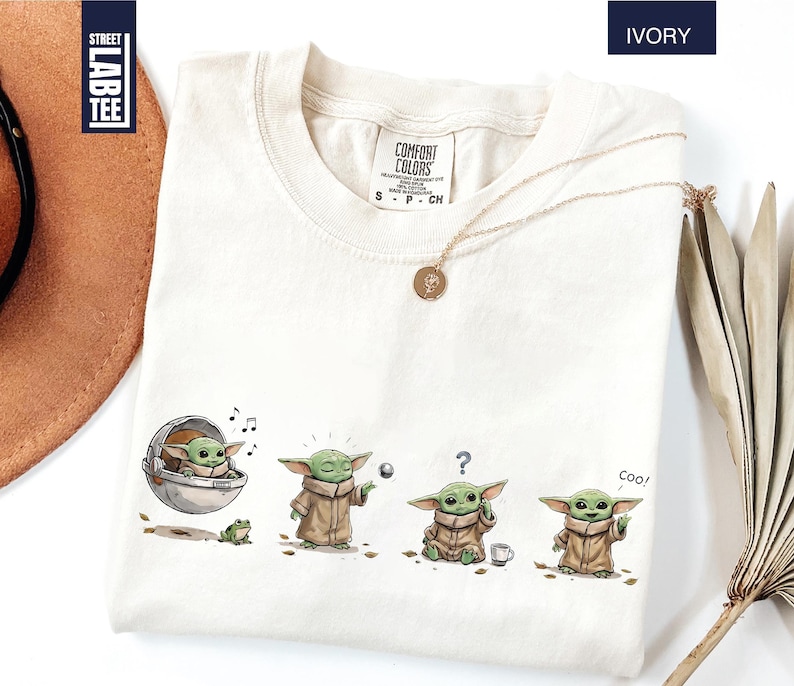 May include: An ivory t-shirt featuring a graphic print of a small green creature in four different poses. The print shows the character in a floating pod, using the force, looking confused with a question mark, and waving with the text Coo.