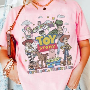 May include: Pink t-shirt featuring a colorful graphic of characters from the animated film "Toy Story." The design includes Woody, Buzz Lightyear, and other characters, with the text "TOY STORY" and "YOU'VE GOT A FRIEND IN ME."