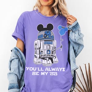 May include: A lilac-colored t-shirt featuring a mashup of R2-D2 and Mickey Mouse. The droid holds a blue balloon. The text "YOU'LL ALWAYS BE MY BB" is printed below the graphic. The shirt has short sleeves and a relaxed fit.