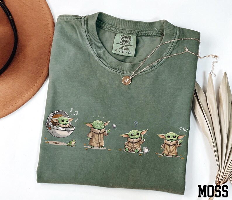May include: A moss green t-shirt featuring a graphic print of a small green creature in four different poses, including sitting in a pod, using the force, looking confused, and waving with the text Coo, with a Comfort Colors brand label inside.