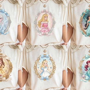 K&ouml;nnte beinhalten: Eine Kollektion von sechs cremefarbenen T-Shirts, die jeweils eine andere Zeichentrickfigur in einem verzierten, mit Schleifen versehenen ovalen Rahmen zeigen. Die Figuren umfassen Elsa, Aurora, Jasmine, Belle, Alice und Ariel in einem weichen Illustrationsstil.