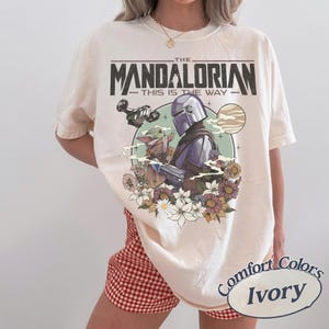 Puede incluir: Una camiseta de manga corta color marfil con un estampado gr&aacute;fico de The Mandalorian y Grogu. El dise&ntilde;o incluye el texto THE MANDALORIAN THIS IS THE WAY, una nave espacial, un planeta y un grupo de flores coloridas en un marco circular.