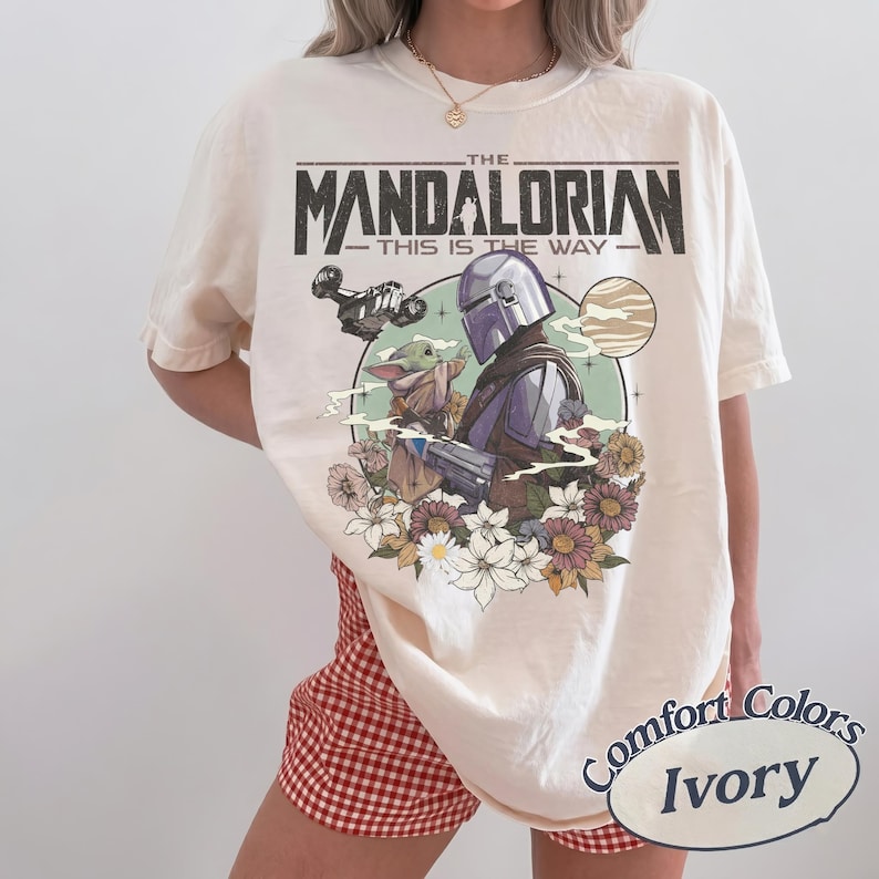 Puede incluir: Una camiseta de manga corta color marfil con un estampado gr&aacute;fico de The Mandalorian y Grogu. El dise&ntilde;o incluye el texto THE MANDALORIAN THIS IS THE WAY, una nave espacial, un planeta y un grupo de flores coloridas en un marco circular.