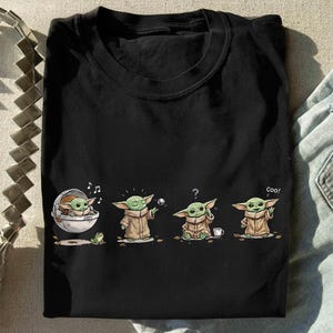 May include: A black short-sleeved t-shirt featuring a graphic print of a small green creature in a tan robe. The illustration shows the character in four poses: sitting in a floating pod, reaching for a ball, looking confused, and saying coo.